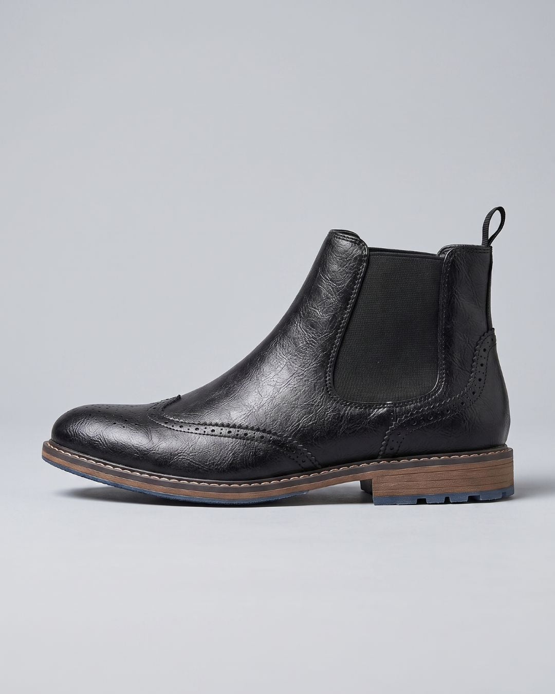 A black leather Chelsea boot with a textured finish, featuring elastic side panels, a pull tab, and a stacked wood-look heel, shown from a side profile on a light gray background.