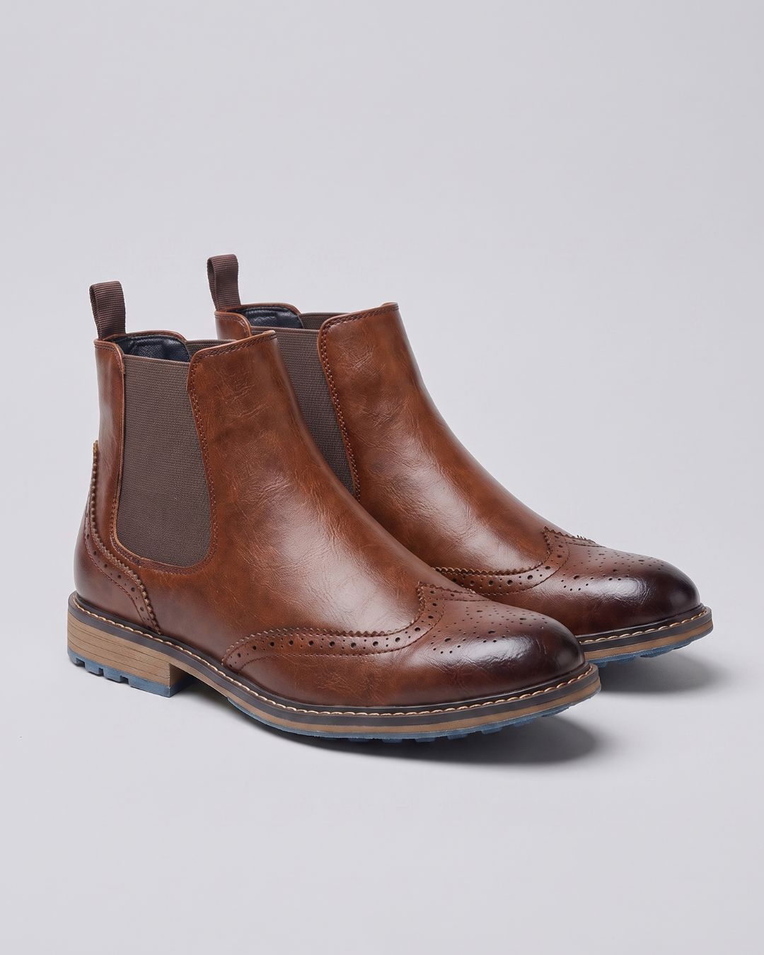 A pair of brown leather Chelsea boots with brogue detailing and elastic side panels, photographed on a light gray studio background.