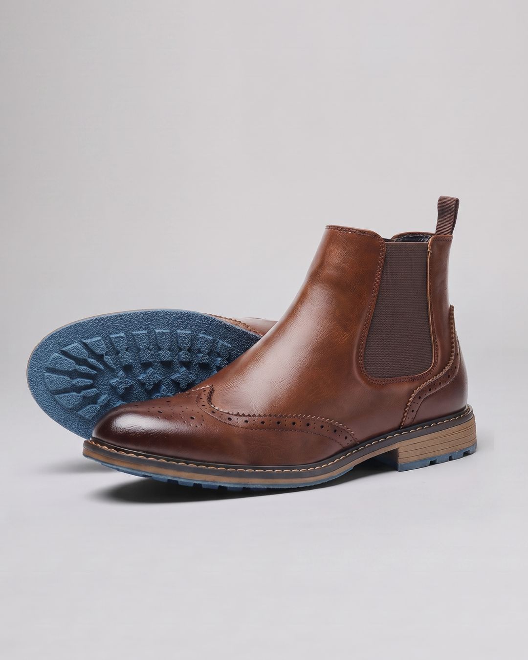 A pair of men's brown leather Chelsea boots with brogue detailing, shown on a light gray background with one boot standing upright and the other showing its blue textured sole.
