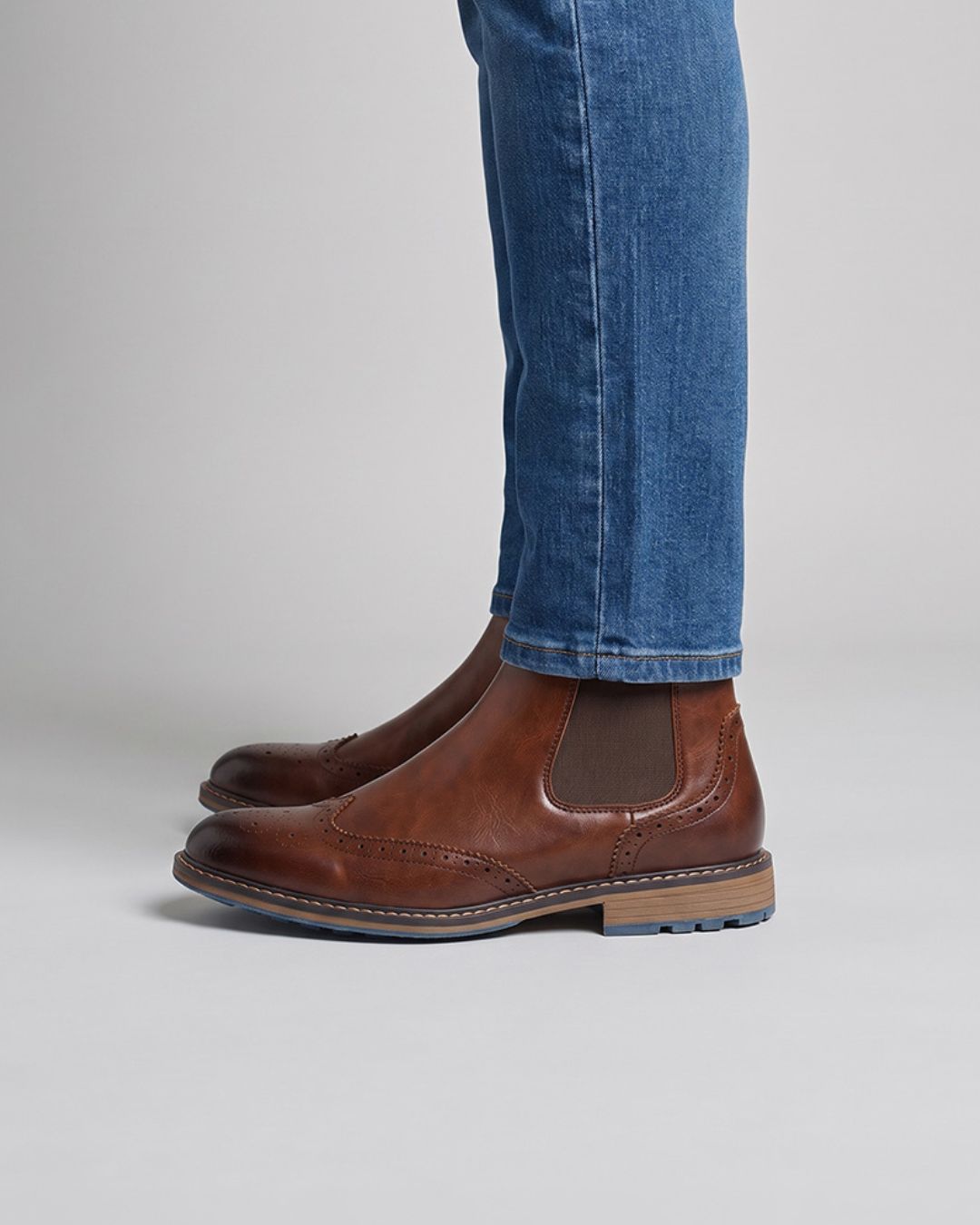 A pair of men's brown leather Chelsea boots with elastic side panels, shown on a model wearing blue jeans against a light gray studio background.