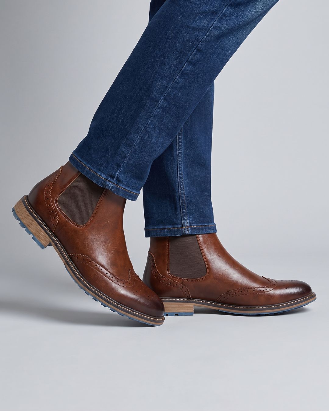 A pair of men's brown leather Chelsea boots with wingtip brogue detailing and dark elastic side panels, shown on a model wearing blue jeans against a light gray studio background.