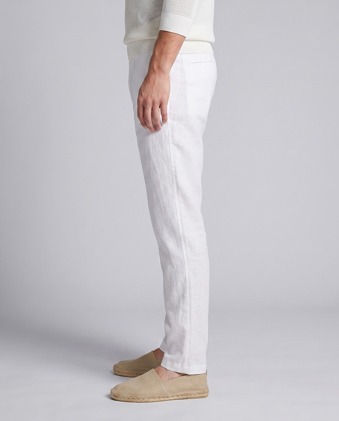A side profile of a person wearing white linen trousers, a cream ribbed knit sweater, and beige espadrille slip-on shoes against a light gray studio background.