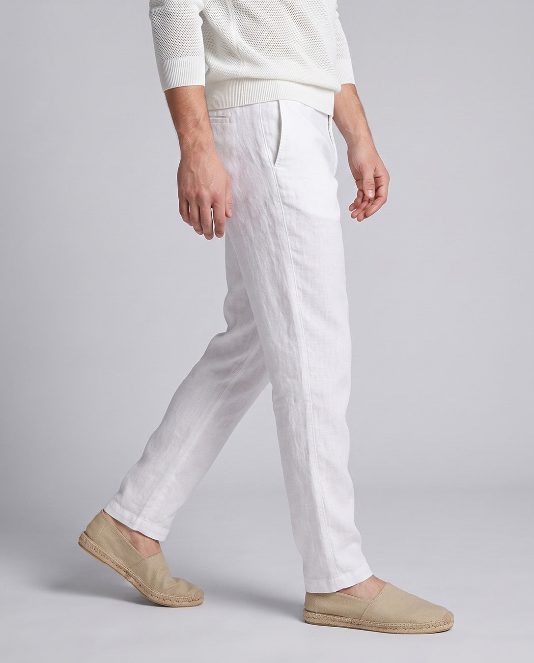A person wearing white linen trousers, a cream knit sweater, and beige espadrille slip-on shoes, shown from the chest down in a side profile against a light gray studio background.