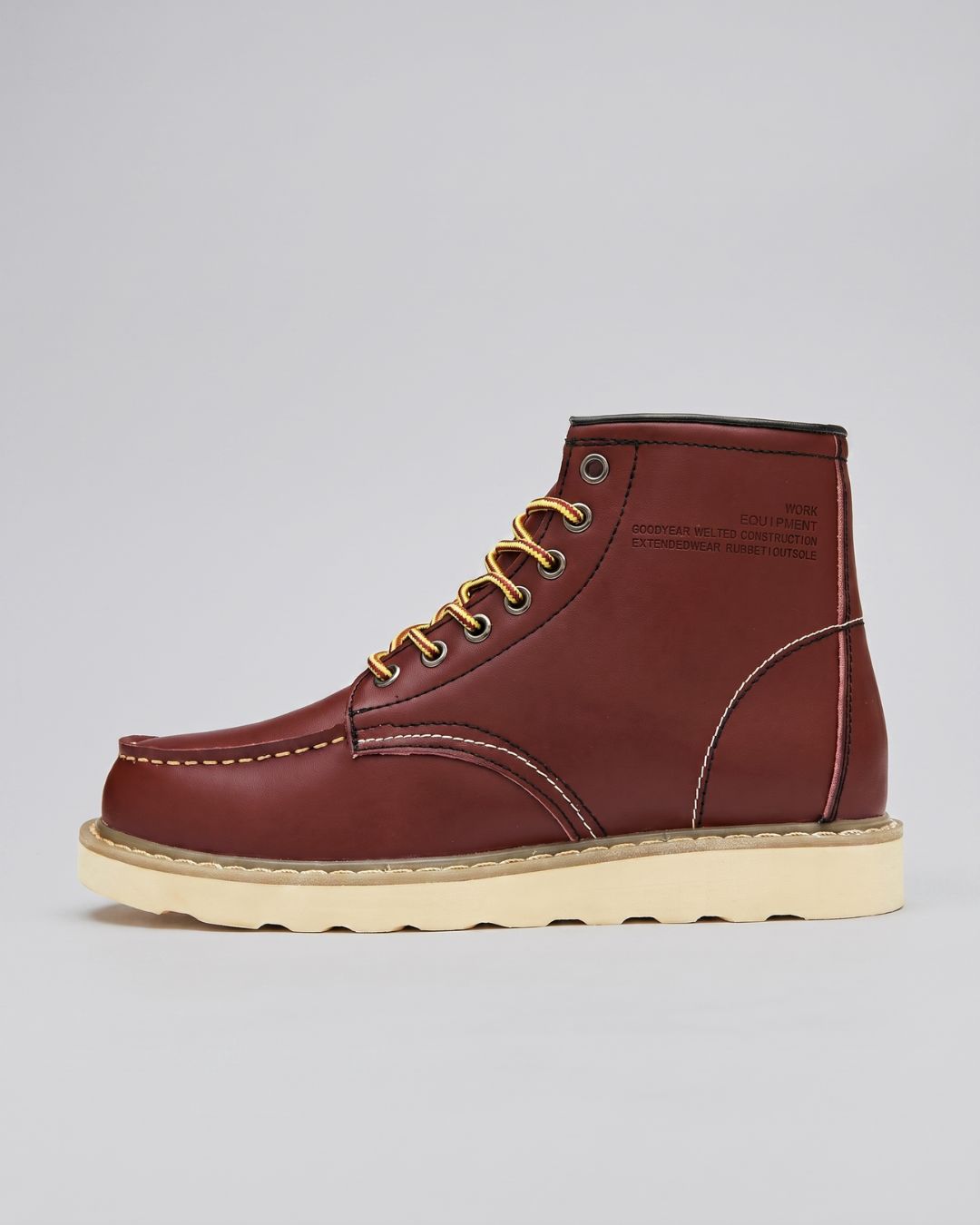 A single mid-cut leather boot in brown red, featuring gold laces threaded through brass eyelets, a cream-colored sole, and subtle contrast stitching on a light gray background.
