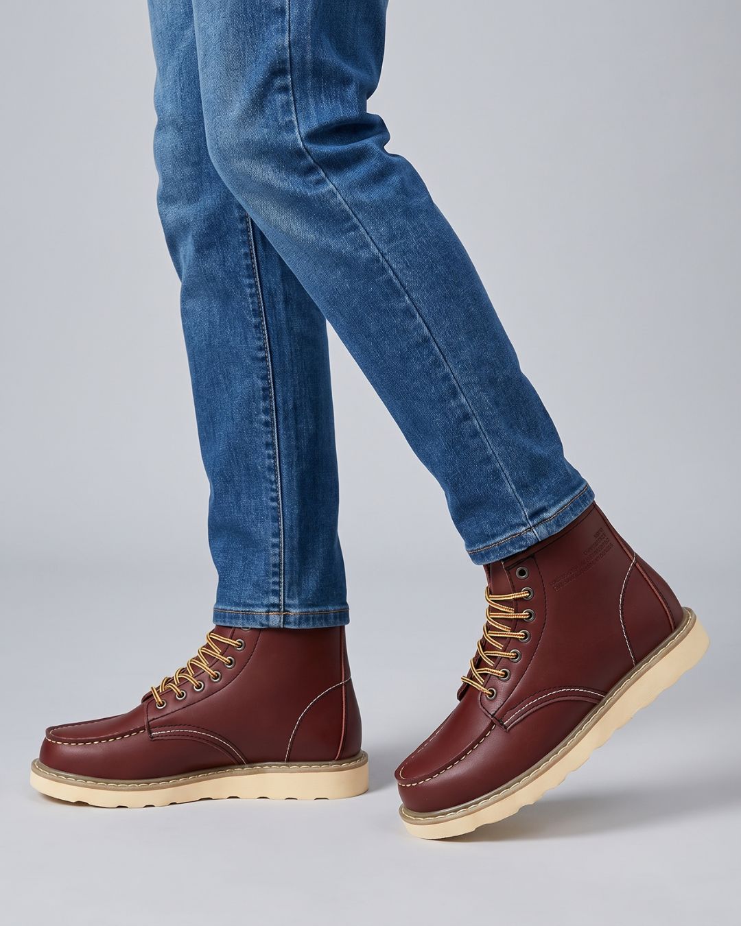 A person wearing blue jeans and brown burgundy lace-up boots with beige soles, shown from the knees down against a light gray studio background.