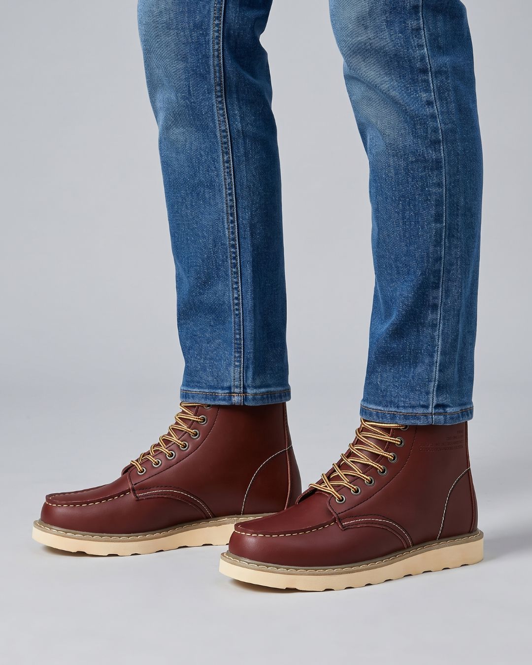 A pair of men's brown leather lace-up boots with tan laces and beige soles, shown on a model wearing blue jeans against a light gray studio background.