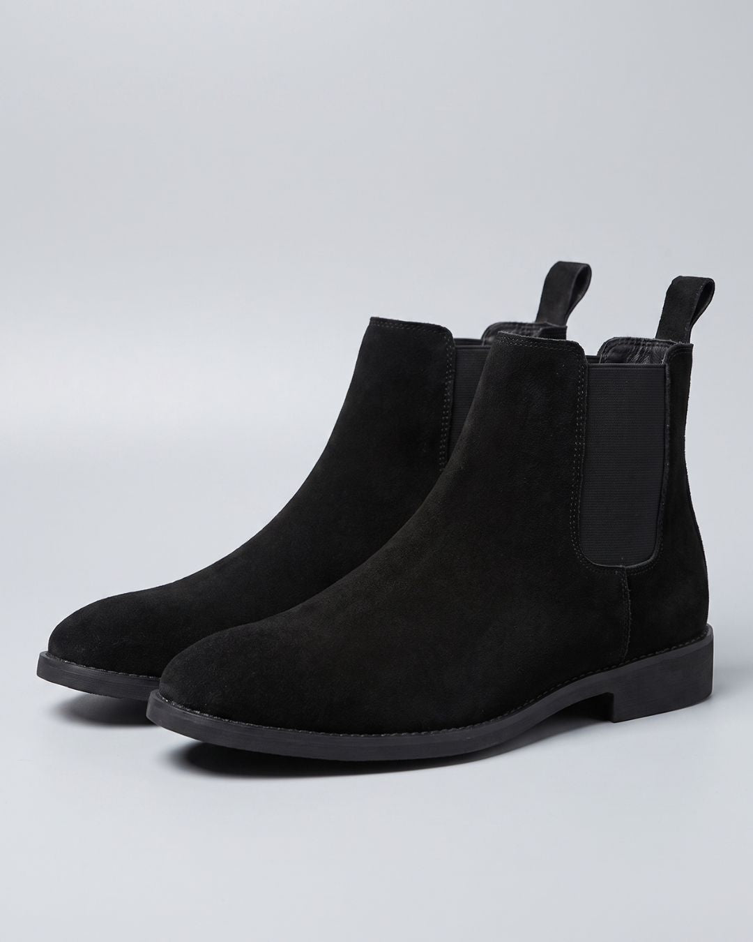 A pair of black suede Chelsea boots with black elastic side panels and pull tabs, shown on a light gray studio background.