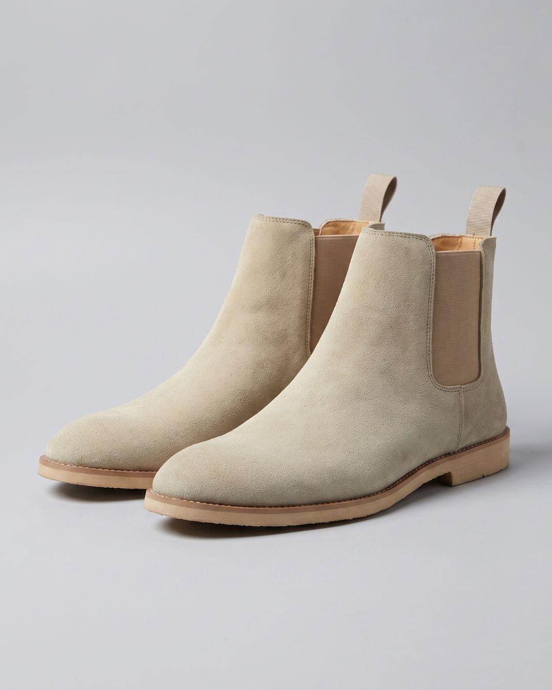A pair of beige suede Chelsea boots with elastic side panels and pull tabs, shown on a light gray studio background.