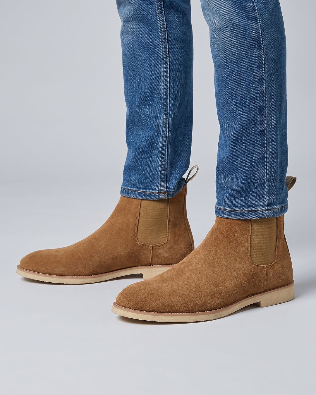 A pair of men's brown suede Chelsea boots worn with blue jeans, shown from a side profile against a light gray studio background.
