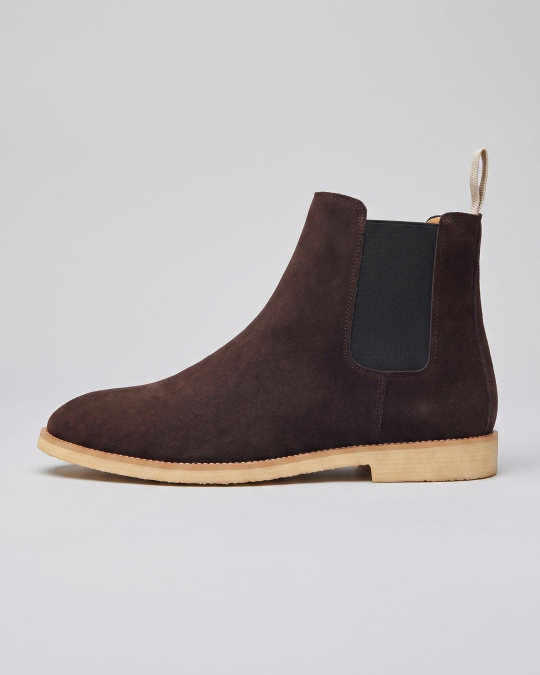 A dark brown suede Chelsea boot with a black elastic side panel and a light tan sole, shown in profile against a neutral light gray background.