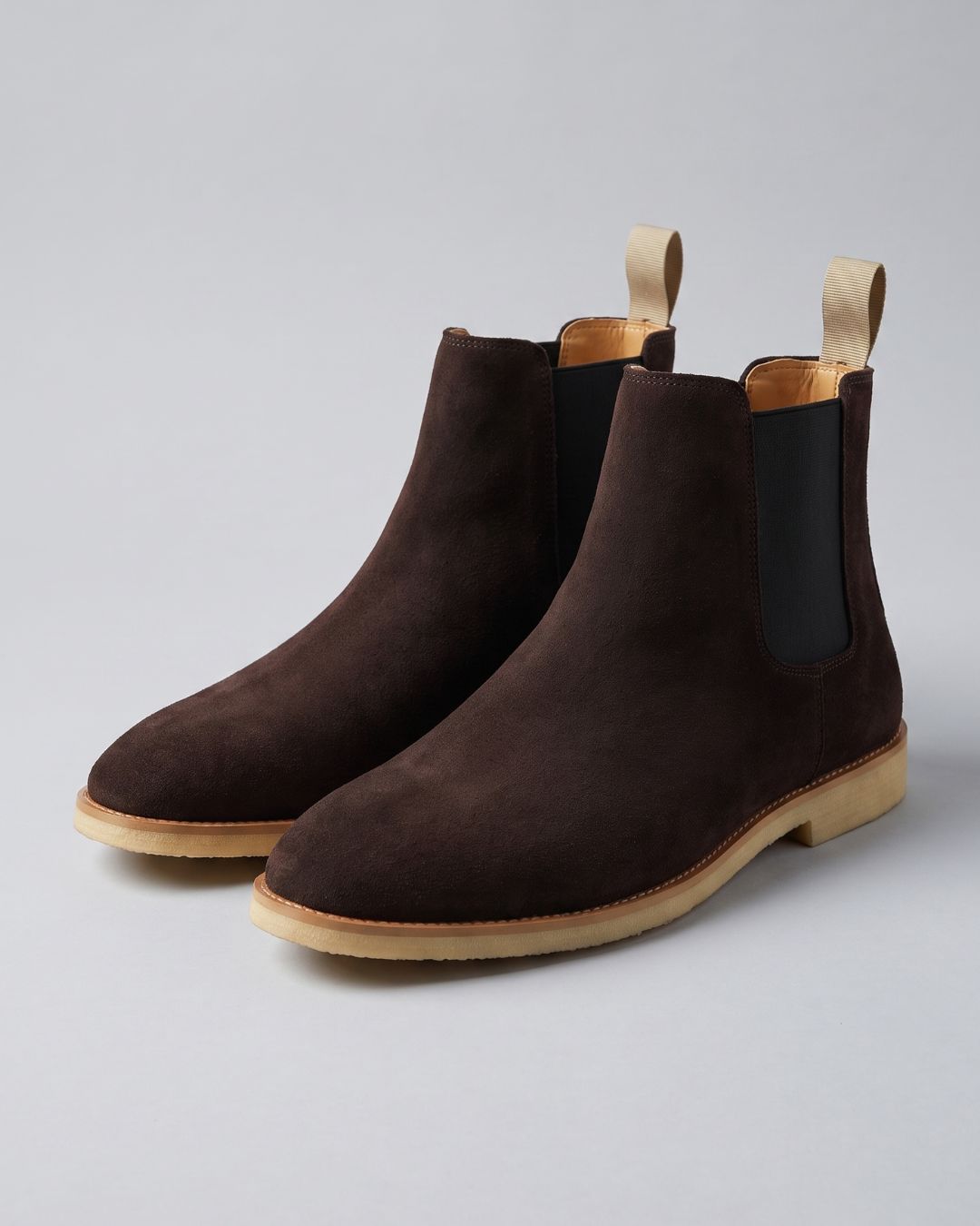 A pair of dark brown suede Chelsea boots with black elastic side panels and light tan pull tabs and soles, photographed on a neutral light gray background.