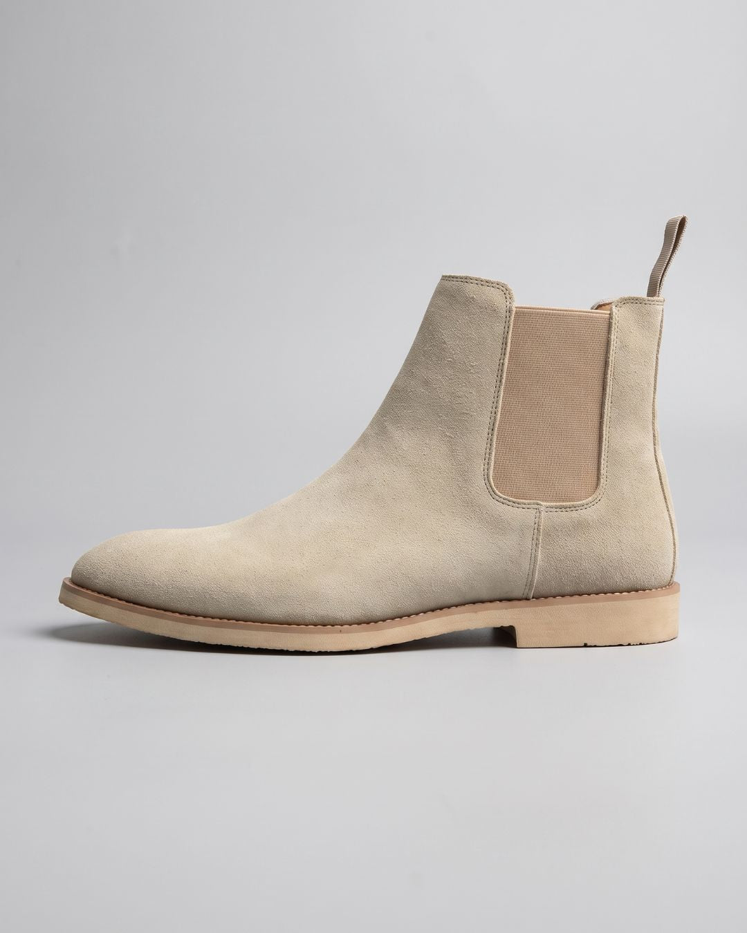 A light beige suede Chelsea boot with a tan elastic side panel and a low stacked heel, shown in profile against a neutral gray background.