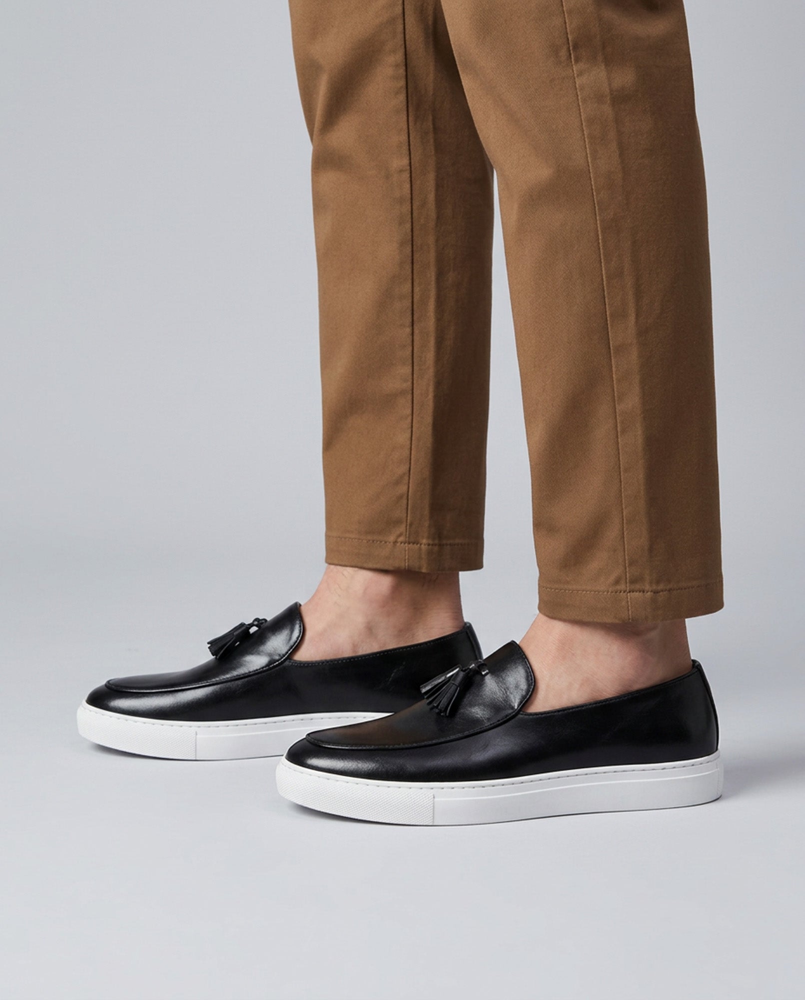 Capri men's black leather tassel loafers styled with tan chino trousers on grey background