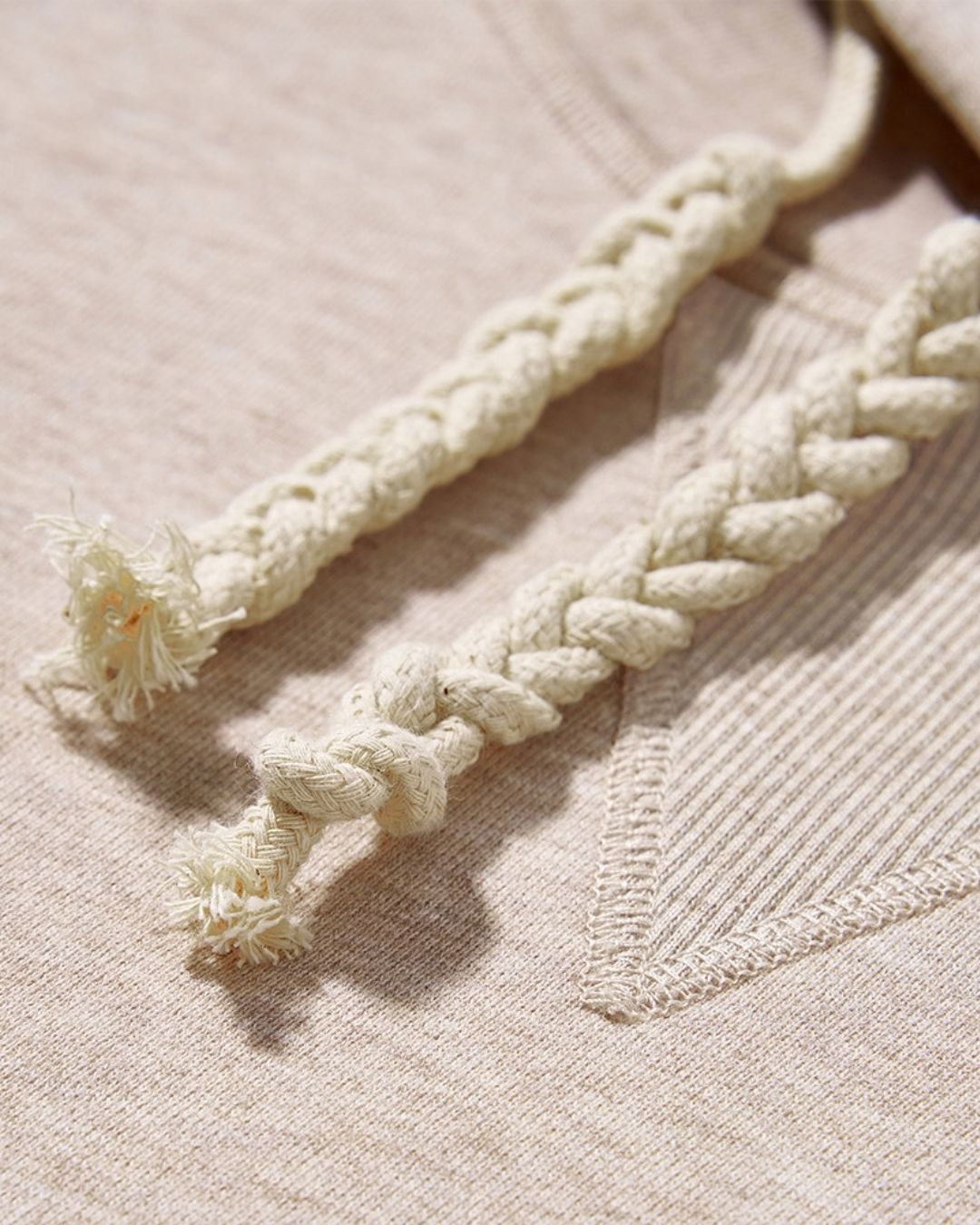 Close-up of a beige cotton hoodie featuring thick, braided drawstring cords and detailed stitching at the neckline.