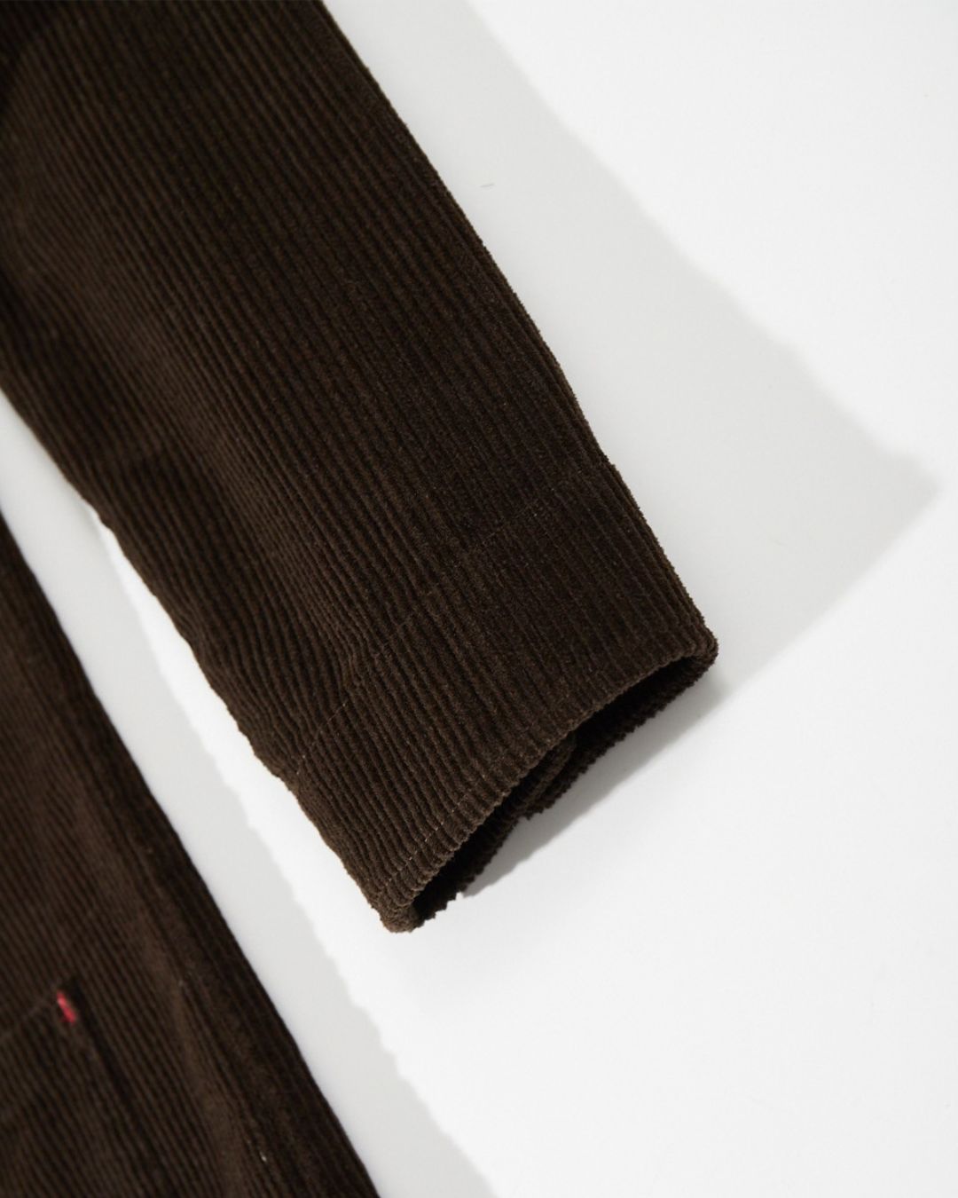 Close-up of a brown ribbed cotton corduroy chore jacket sleeve with a button cuff and subtle red bar-tack stitching.