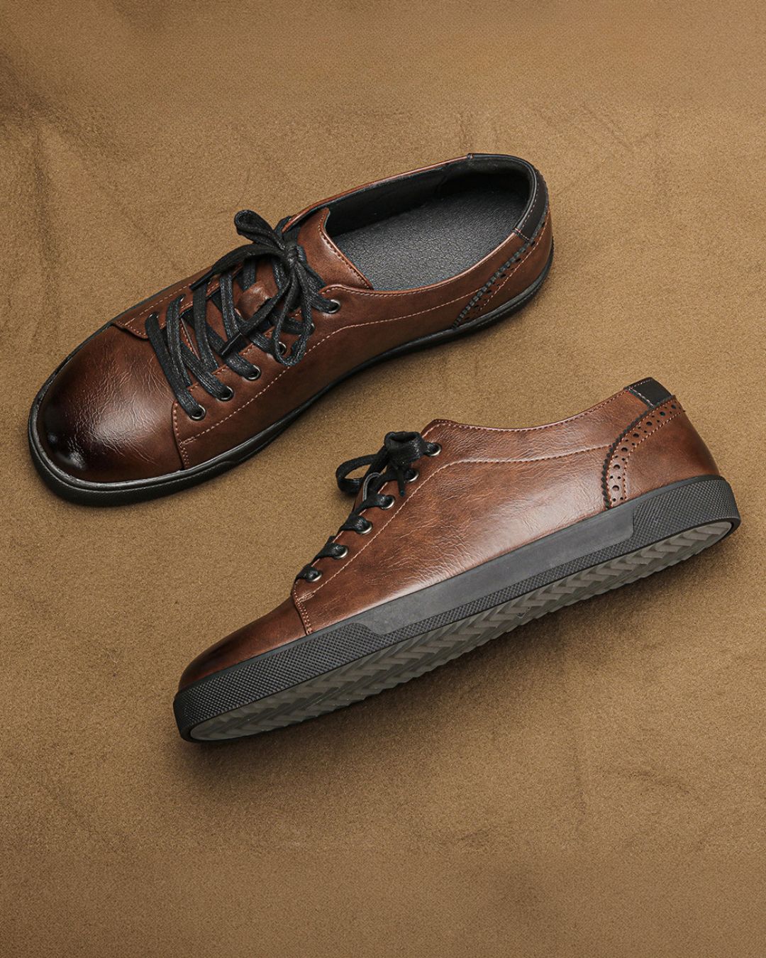 Men’s brown low-top dress sneakers with burnished faux leather, black laces, brogue heel detailing, and a flat rubber sole.