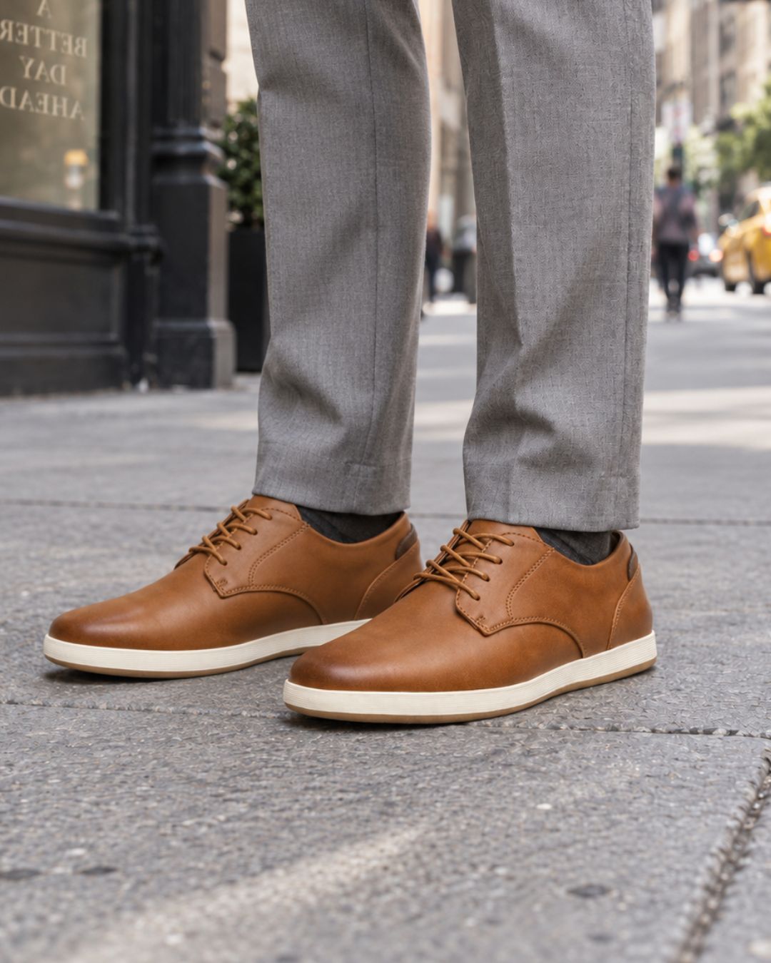 A person wearing brown leather lace-up shoes with off-white soles, paired with gray trousers, standing on a city sidewalk.