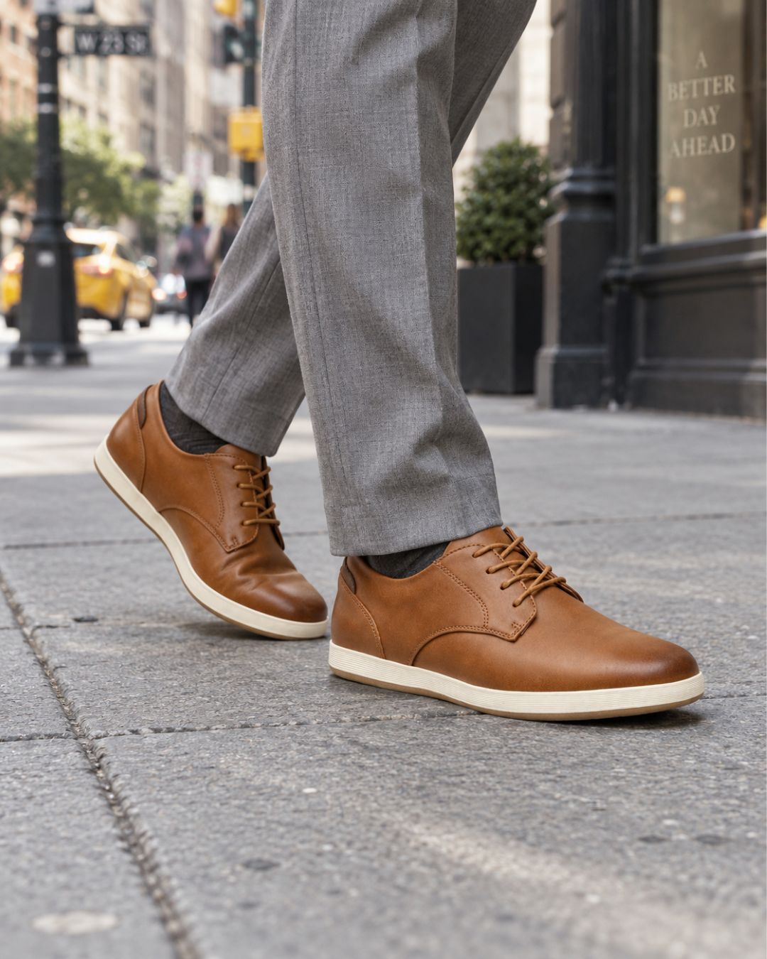 A person wearing brown leather lace-up dress shoes with off-white soles, paired with gray trousers, walking on a city sidewalk.