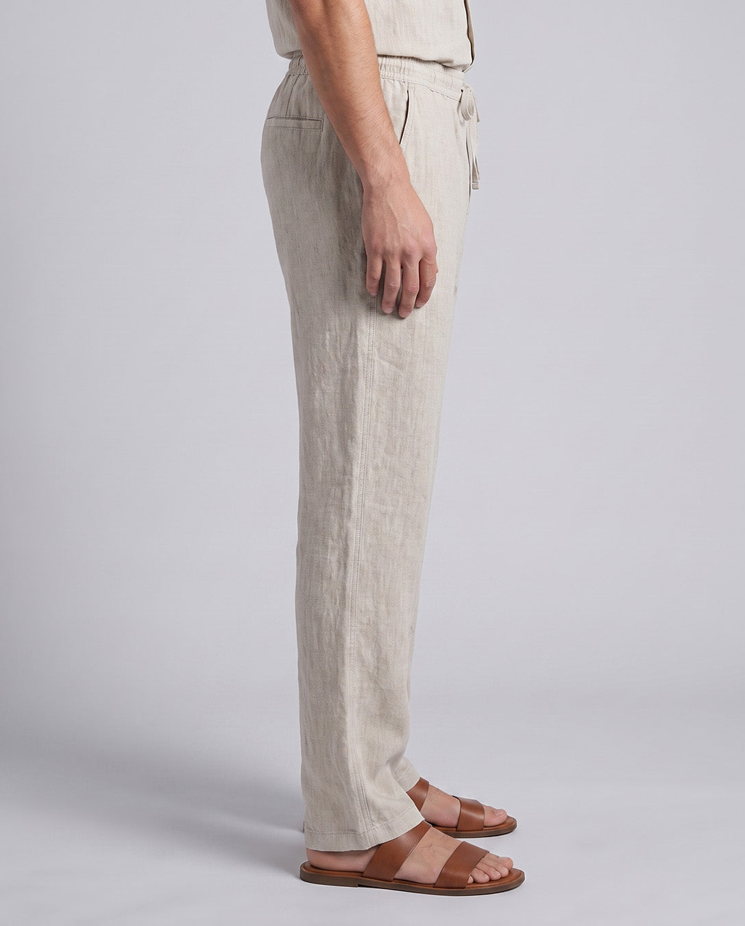 A side view of a person wearing light beige linen drawstring trousers and brown leather slide sandals against a light gray studio background.