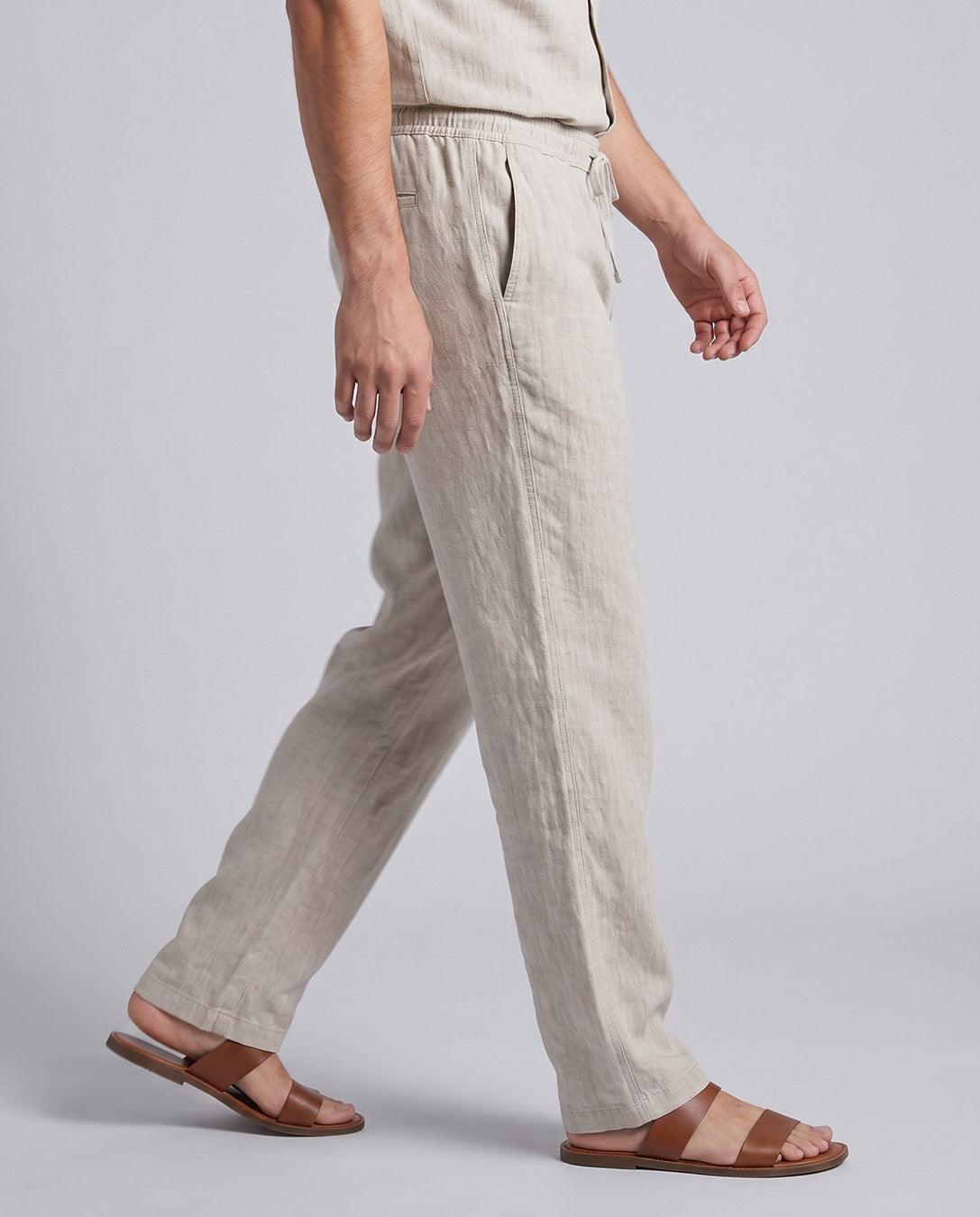 A person wearing light beige linen trousers and brown leather slide sandals, shown from the waist down in a side-profile walking pose against a light gray studio background.