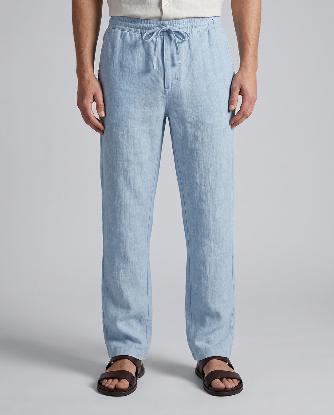 A person wearing light blue drawstring lounge pants with a linen-like texture, paired with dark brown slide sandals against a neutral gray studio background.