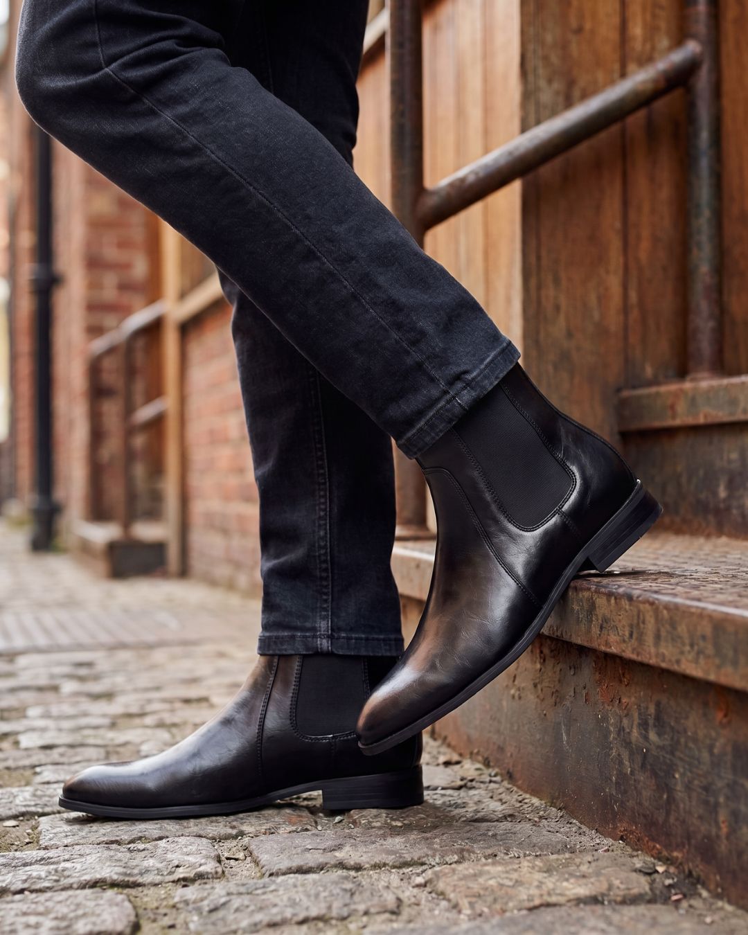 A person wearing black leather Chelsea boots with dark gray jeans, standing on a cobblestone street next to rustic wooden steps.