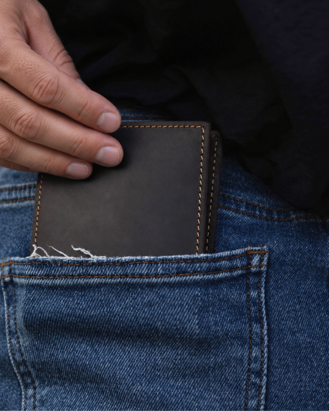 Dark brown Crazy Horse leather bifold wallet with contrast stitching being placed into a blue denim jeans pocket.