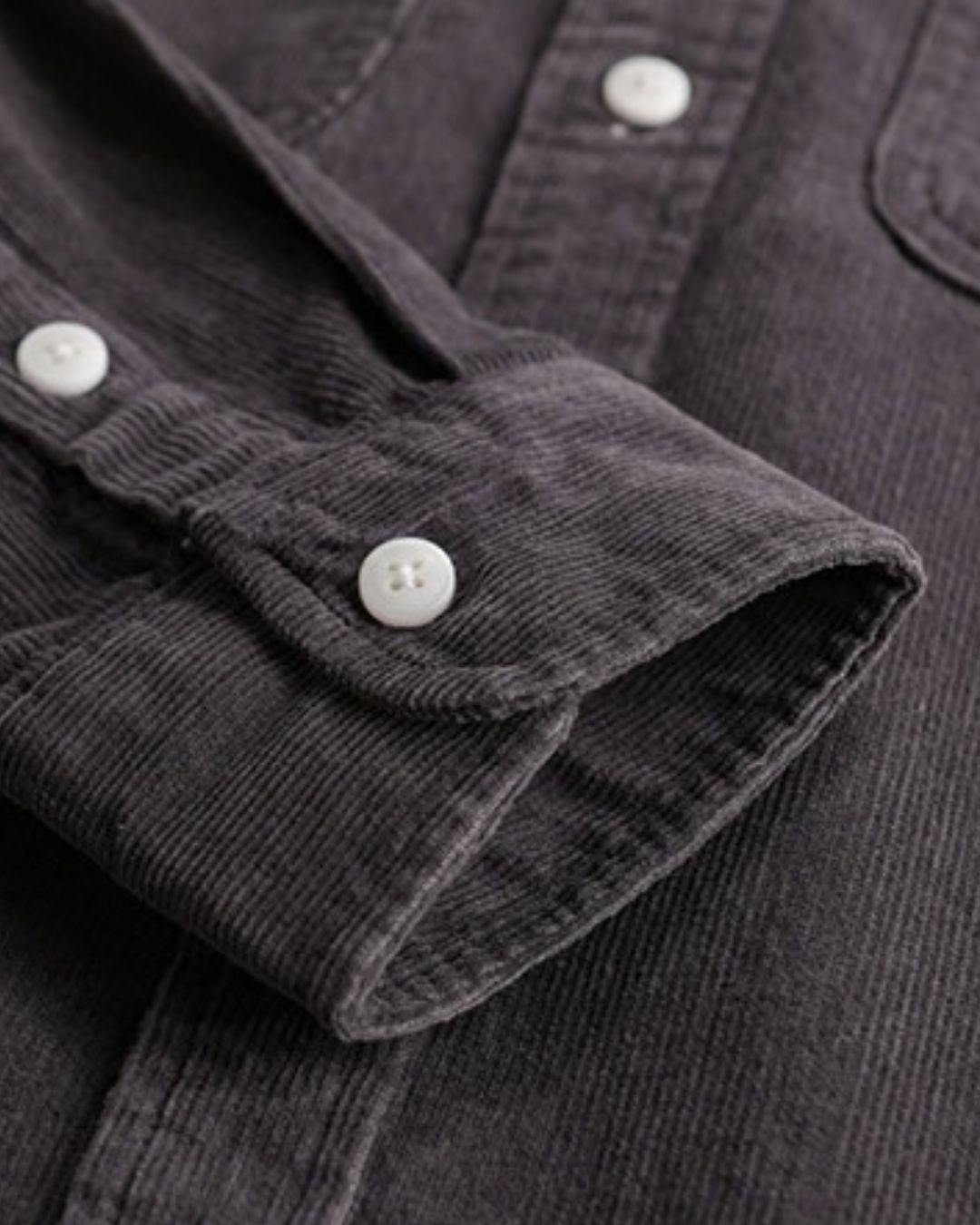 Close-up of a charcoal grey cotton corduroy shirt featuring ribbed texture, long sleeves with button cuffs, and white buttons.