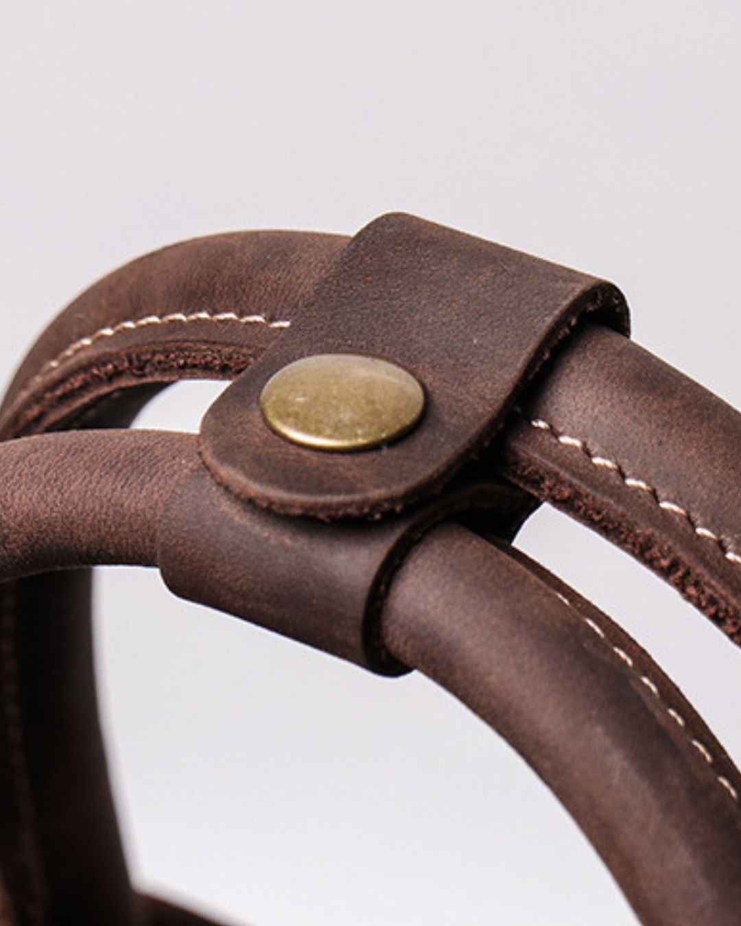 Close-up of a dark brown leather strap with white stitching and a round brass snap closure, attached to a matching brown leather loop against a light gray background.