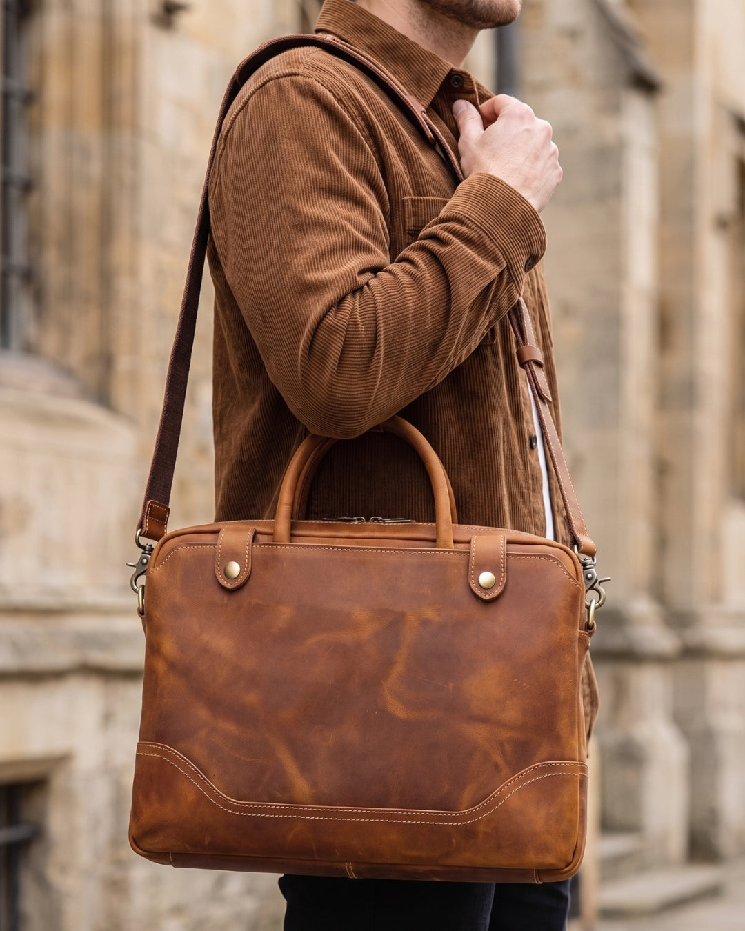A man wearing a brown corduroy shirt carries a cognac brown distressed leather briefcase-style shoulder bag with a top handle and long strap, photographed outdoors against a blurred stone building.