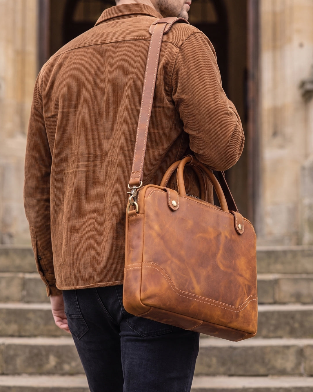 A man wearing a brown corduroy shirt and dark jeans carries a cognac brown distressed leather briefcase-style shoulder bag with a long strap and top handles, standing on stone steps in front of a building.