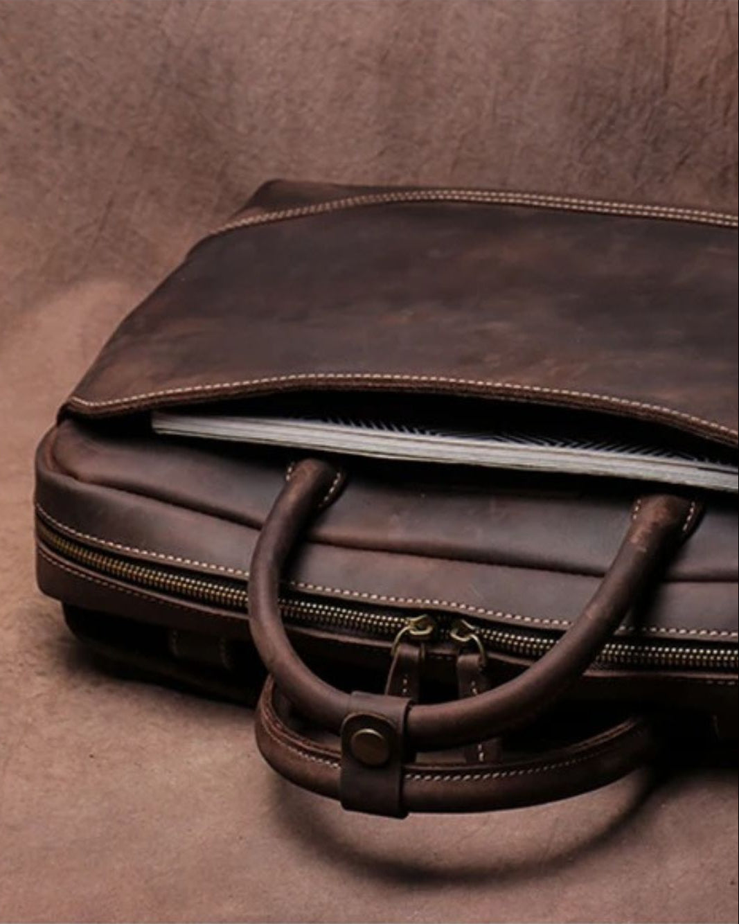 A dark brown distressed leather briefcase or laptop bag with dual top handles, shown open with a notebook inside, set against a matching brown textured background.