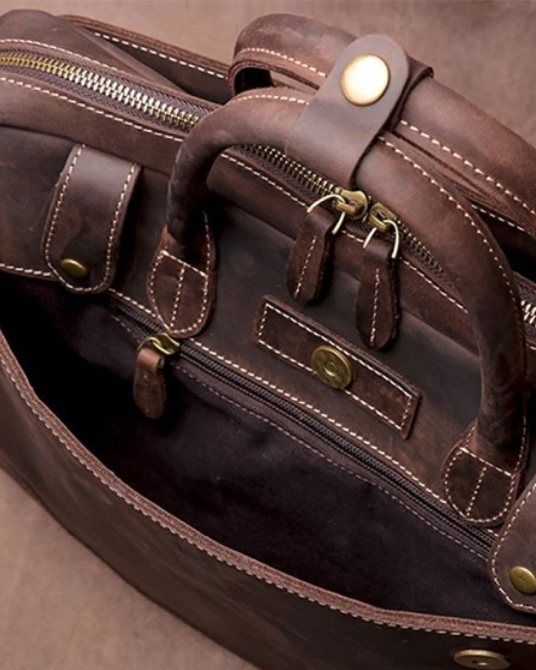 Close-up of a dark brown leather duffel bag with brass hardware, showing the interior zippered compartments, double top handles, and contrast stitching.