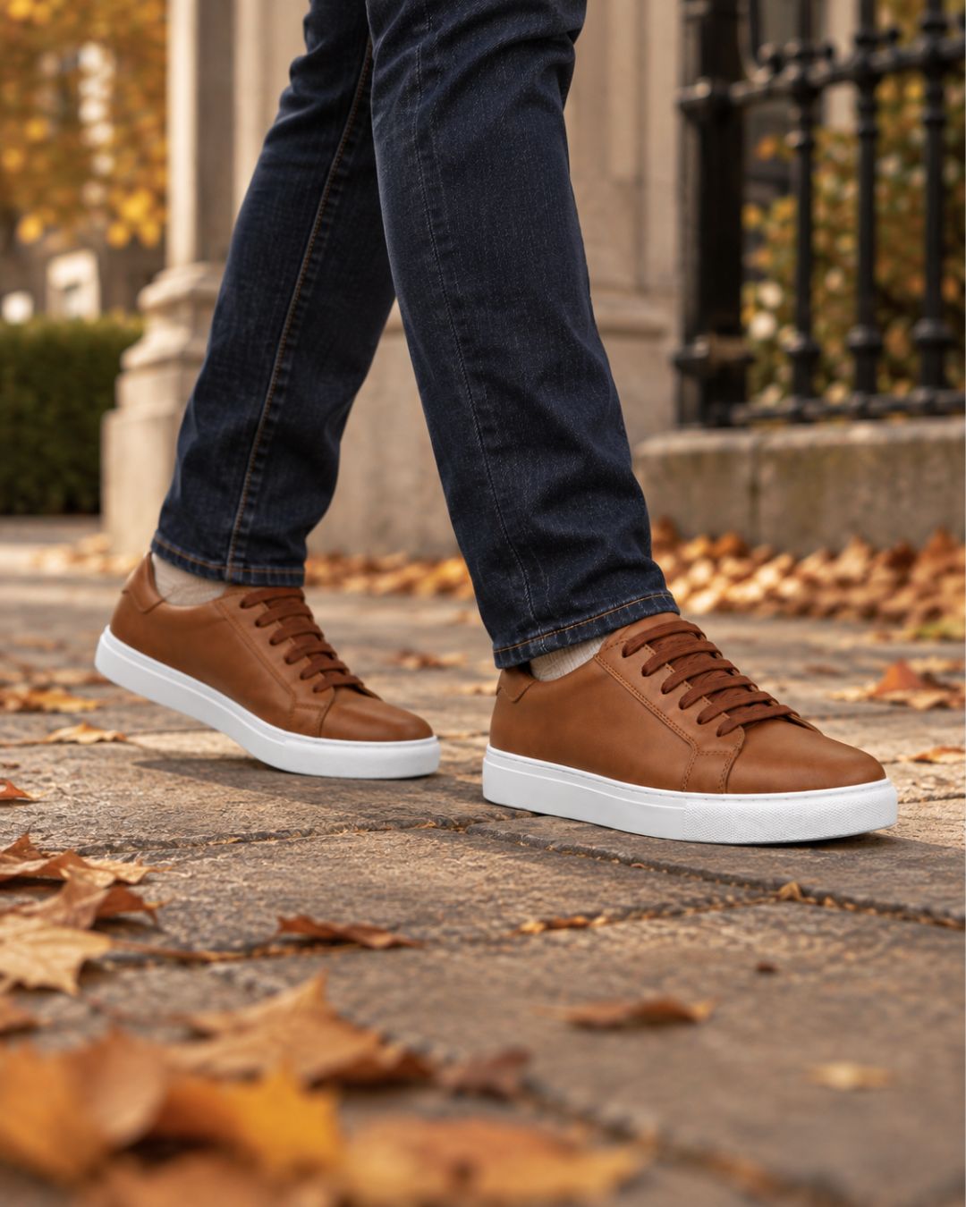 Tan leather low-top dress sneakers with brown laces and a white rubber sole, worn with dark denim jeans on a sidewalk.