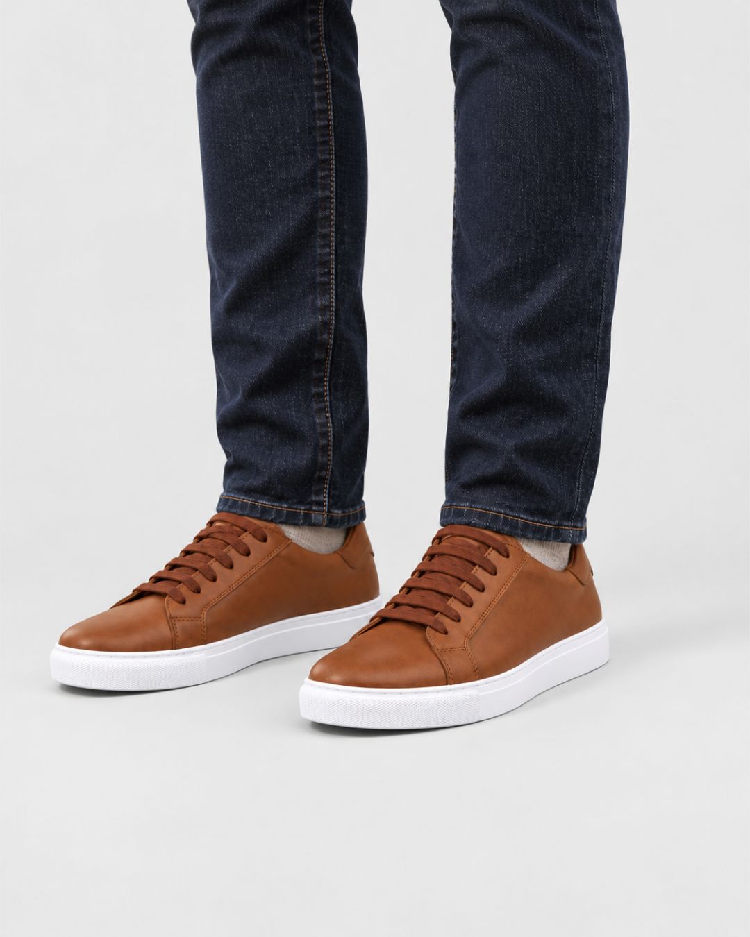 Men’s tan leather low-top dress sneakers with matching laces and a white rubber sole worn with dark denim jeans.
