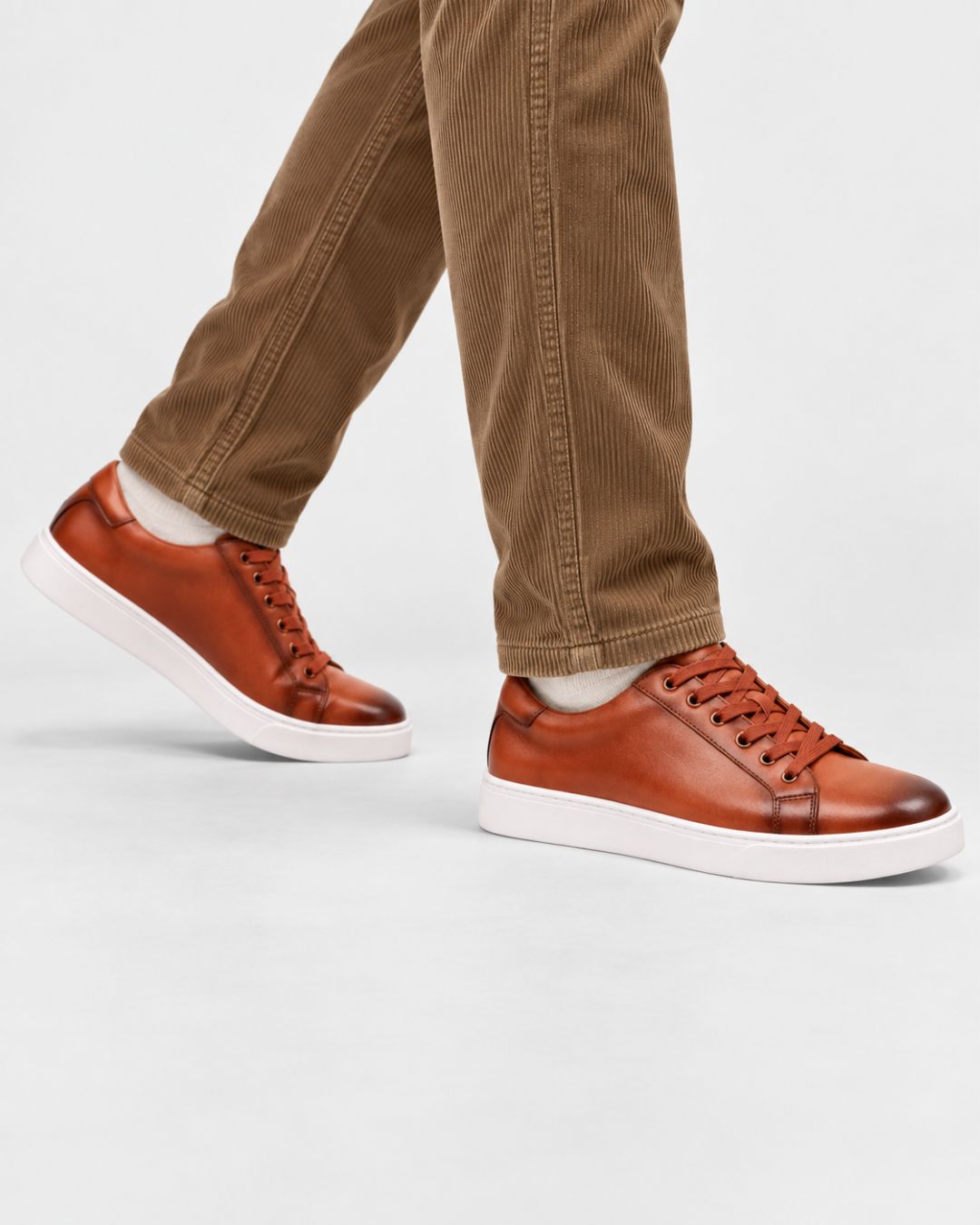 Cognac genuine leather low-top dress sneakers with a white rubber sole, worn with brown corduroy trousers.