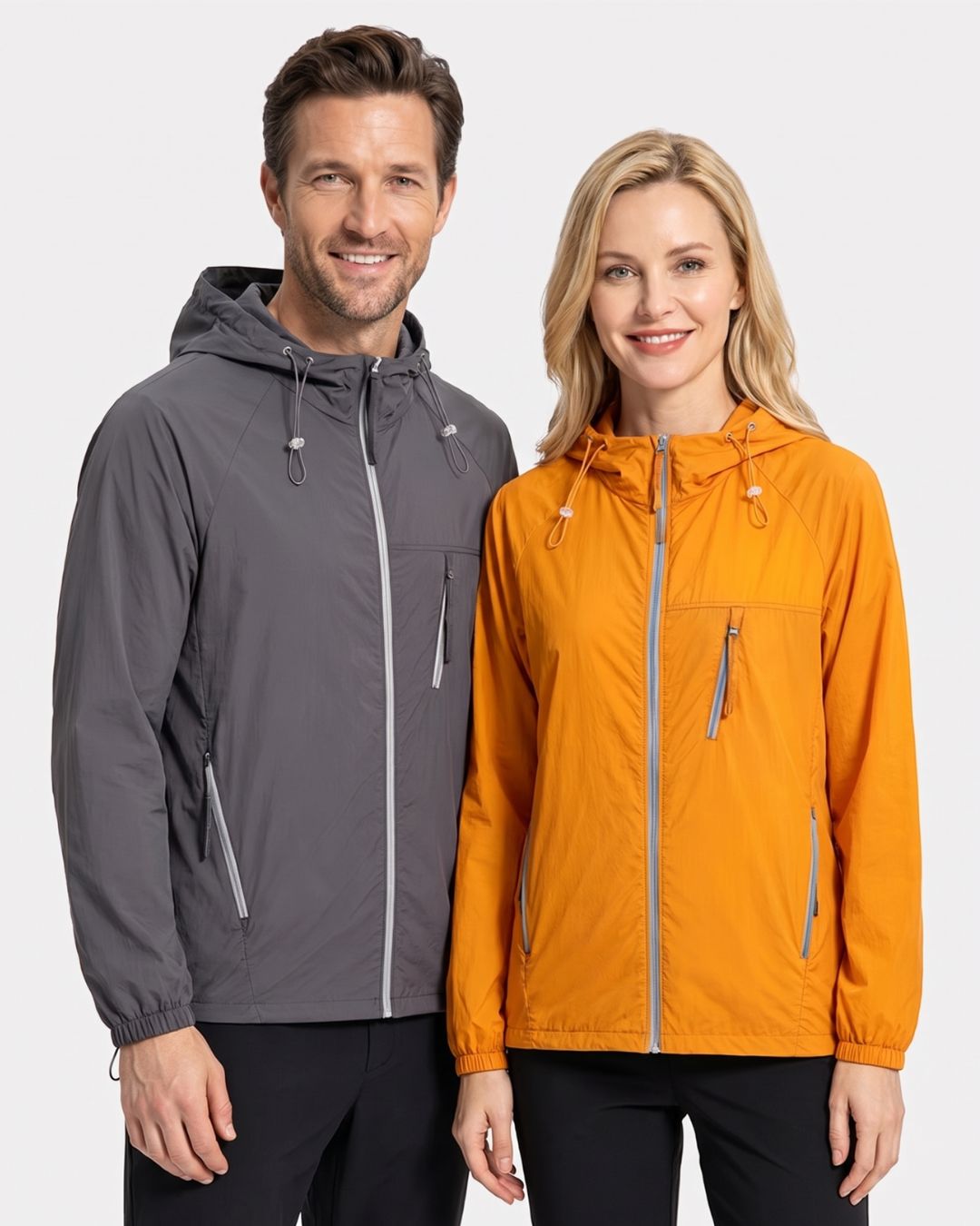 Man and woman wearing lightweight, hooded waterproof windbreakers in charcoal grey and orange with zippered chest pockets.