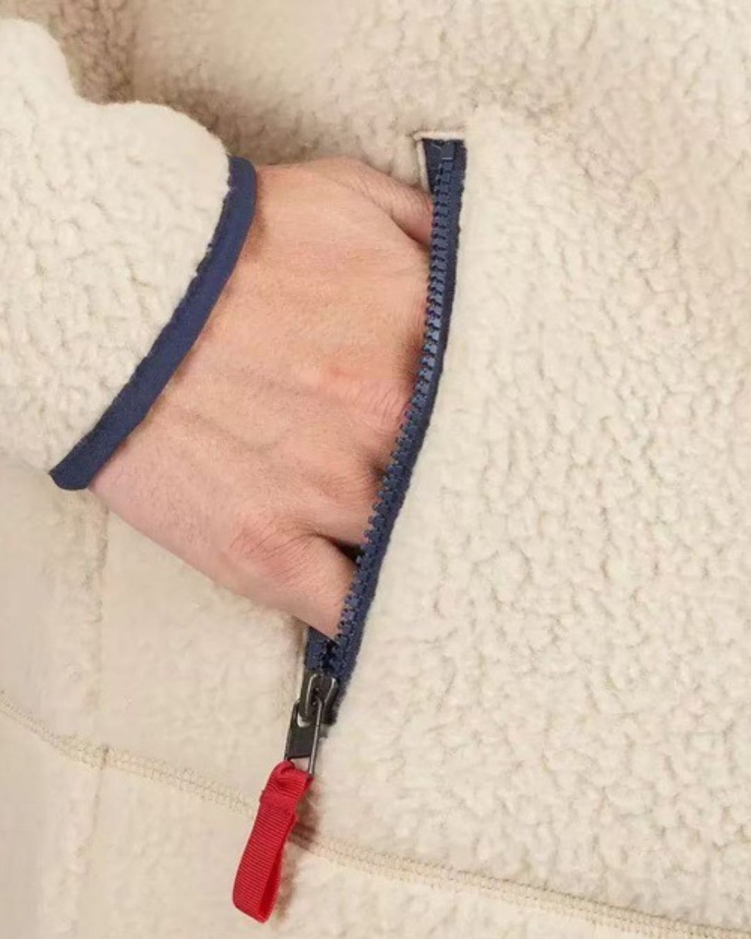 Close-up of a cream teddy sherpa fleece jacket with a navy blue zippered pocket and red pull tab.