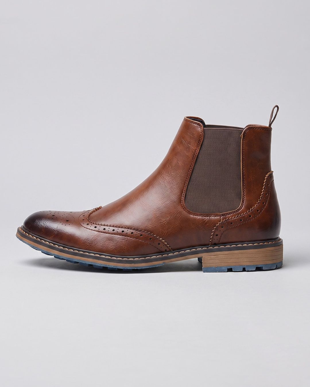 A brown Chelsea boot with wingtip brogue detailing, a dark brown elastic side panel, and a stacked wood-look heel, shown in profile against a light gray background.
