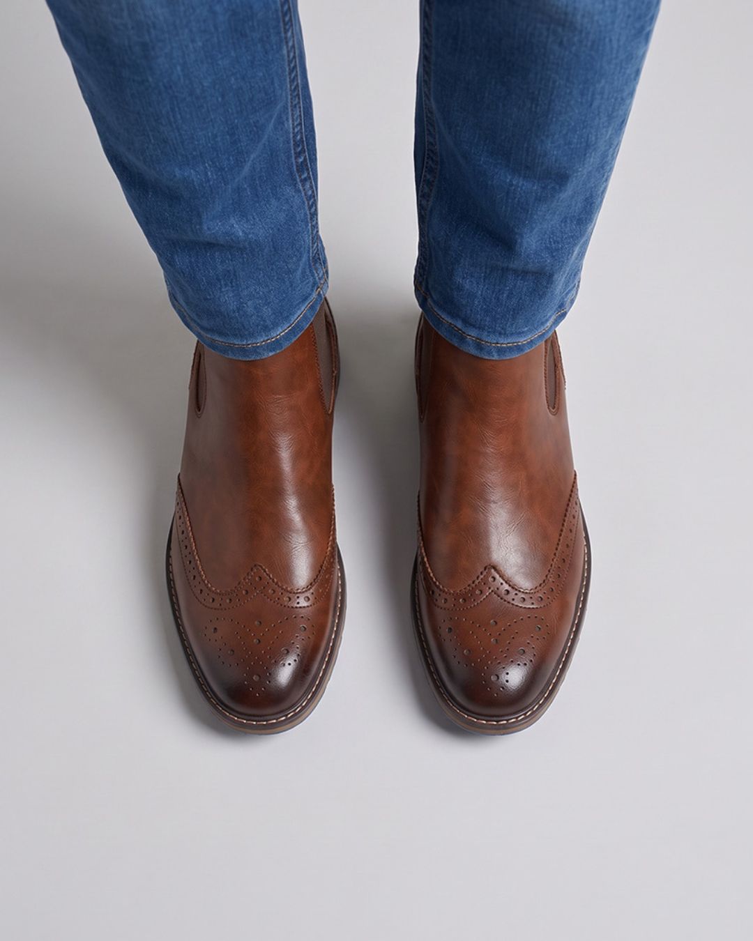 A pair of men's brown leather wingtip brogue boots worn with blue jeans, shown from a top-down view on a light gray background.