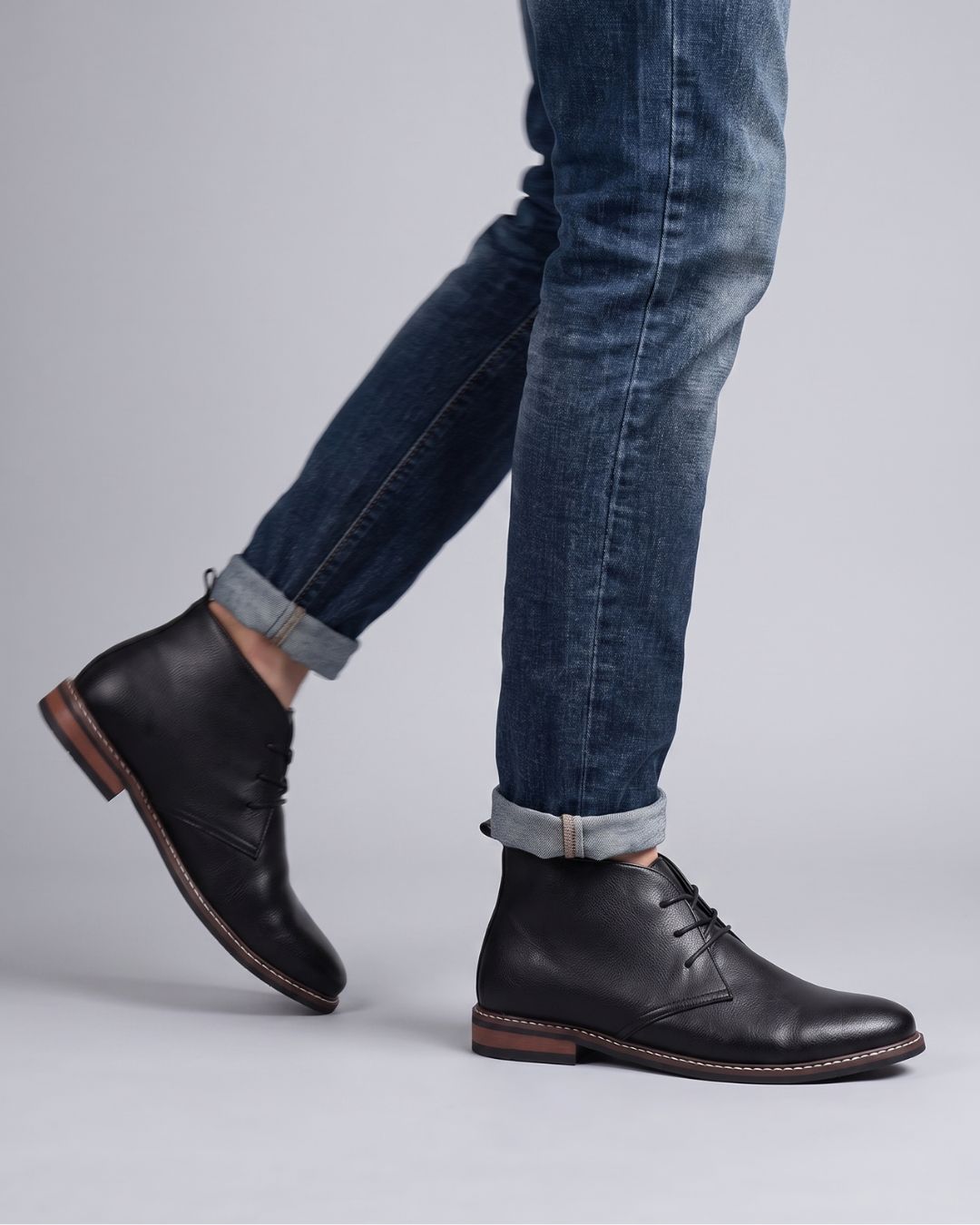 A person wearing dark blue cuffed jeans and black leather lace-up ankle chukka boots, shown from the knees down by Hudson & Tate.
