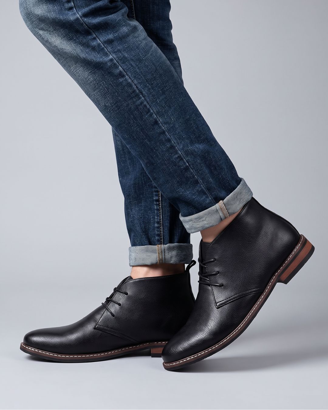 A person wearing dark blue cuffed jeans and black leather lace-up ankle chukka boots, shown from the knees down by Hudson & Tate.