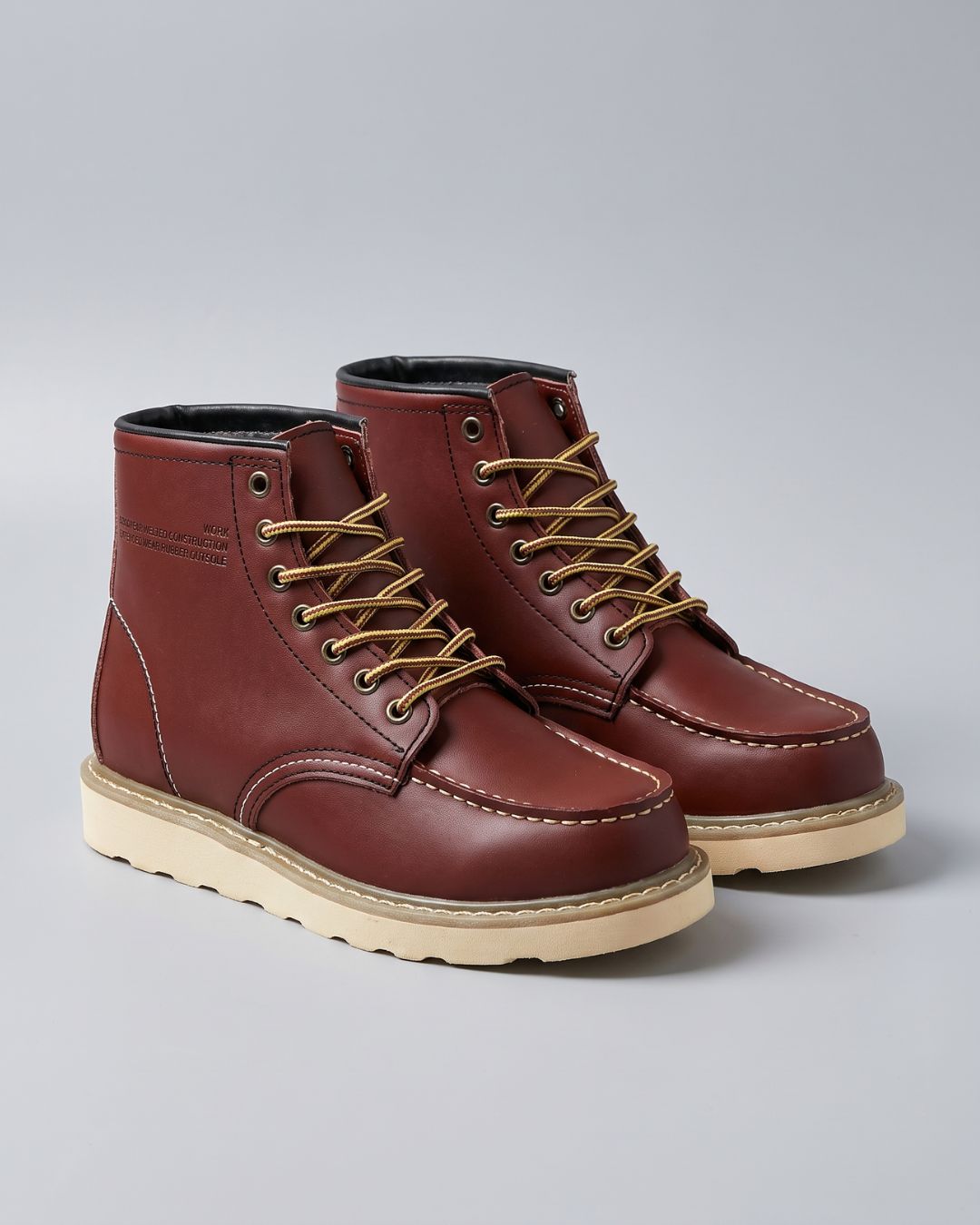 A pair of brown leather lace-up ankle boots with tan laces and beige soles, photographed on a light gray studio background.