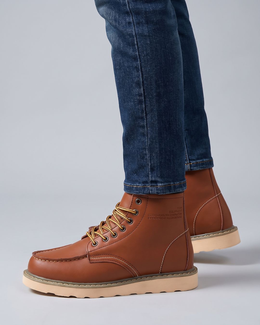 A pair of men's cognac leather lace-up boots with tan laces and light beige soles, shown on a model wearing blue jeans against a light gray studio background.
