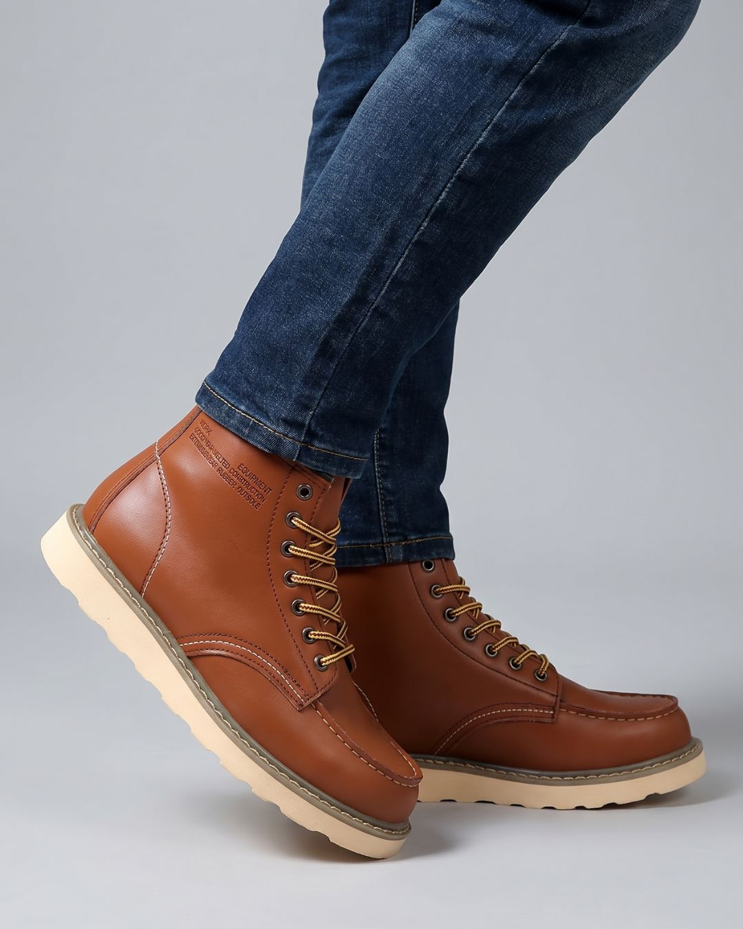 A pair of men's cognac leather lace-up boots with thick beige soles, shown on a model wearing blue jeans against a light gray studio background.