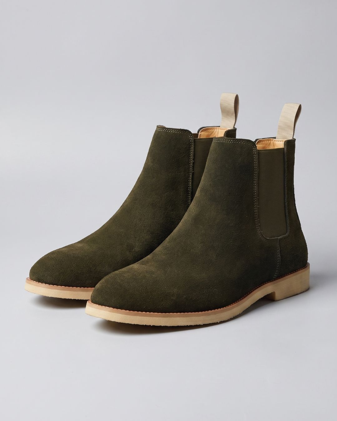 A pair of dark olive green suede Chelsea boots with tan soles and pull tabs, photographed on a neutral light gray background.