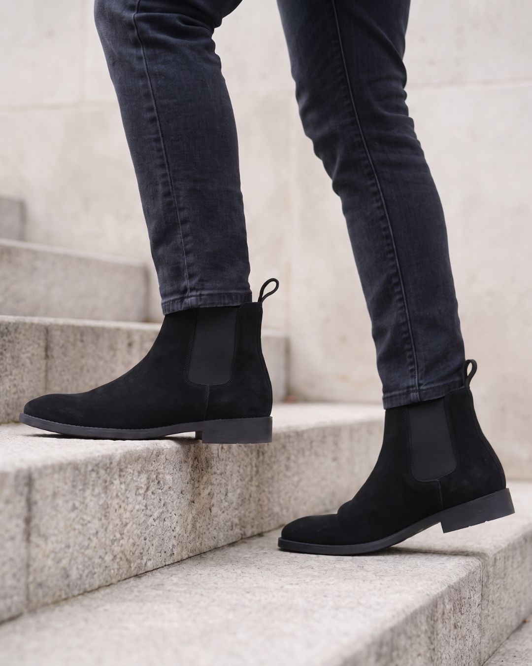 Black suede Chelsea boots worn with dark gray jeans, shown on a person standing on light gray stone steps.