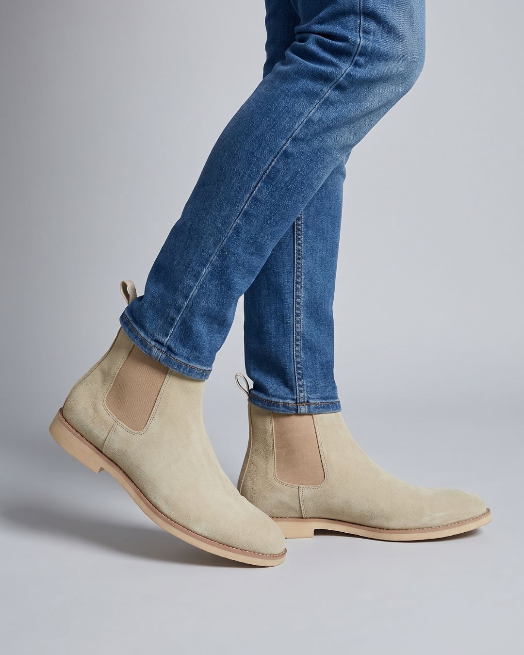 Beige suede Chelsea boots worn with blue jeans, shown from the side against a light gray studio background.