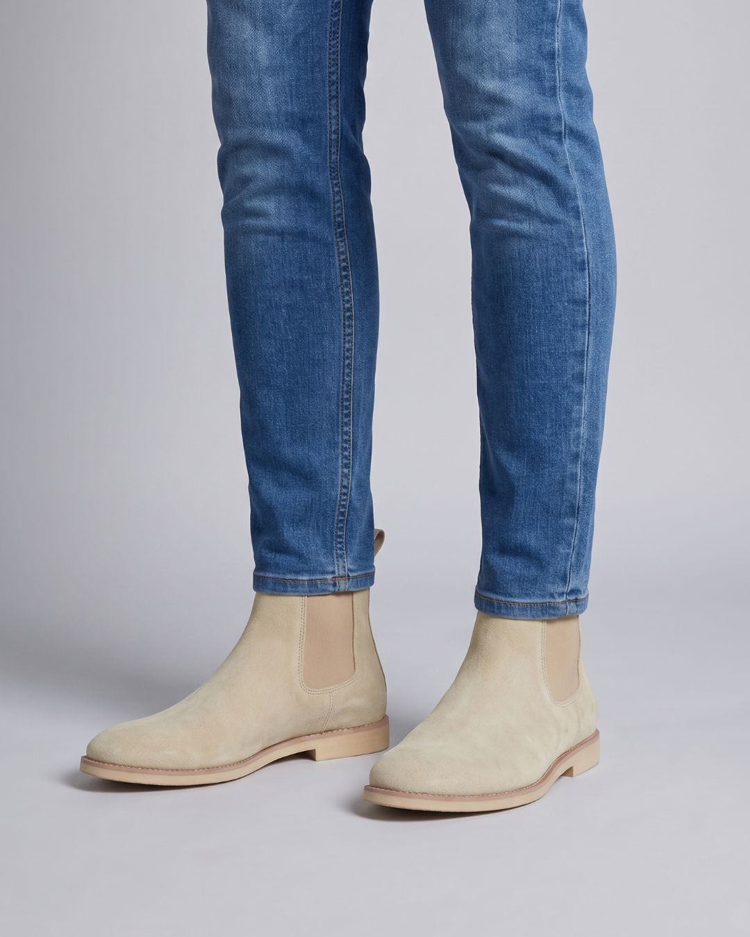 Beige suede Chelsea boots worn with blue jeans against a light gray studio background.