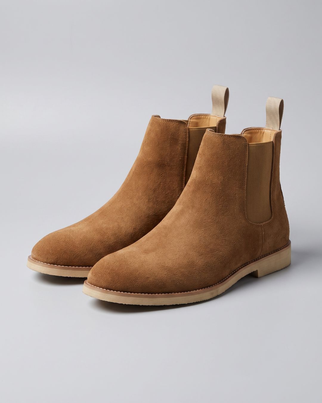 A pair of brown suede Chelsea boots with elastic side panels and pull tabs, shown on a light gray studio background.